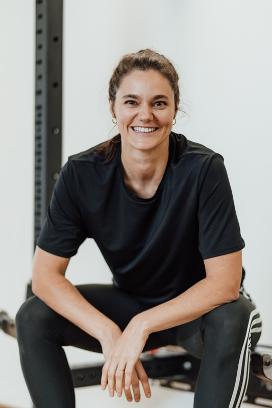 Portrait Michelle Wegener sitzt lachend in schwarzer Kleidung auf einem Rack